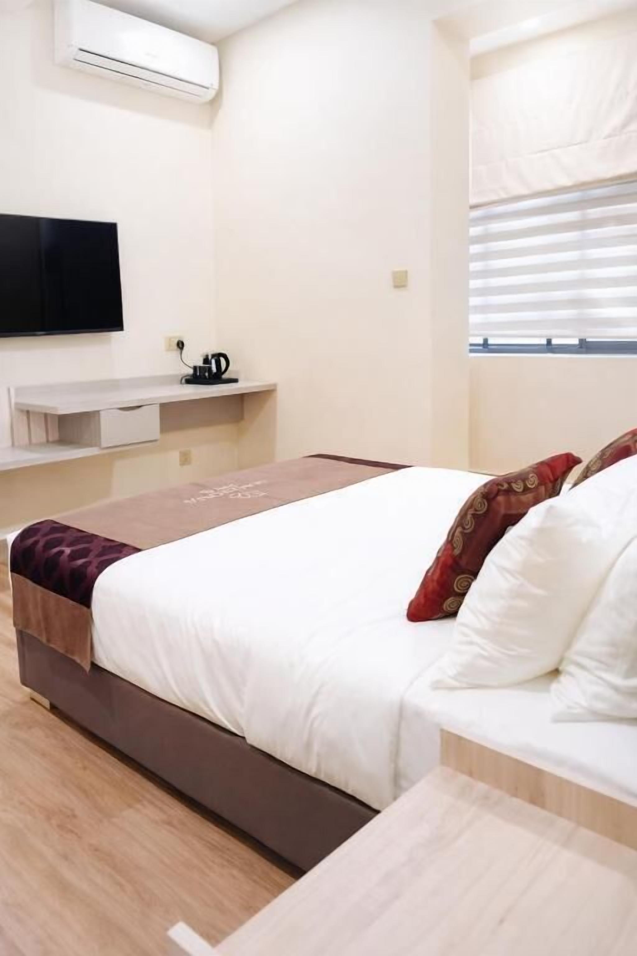 A hotel room with a bed, a bedside table, a flat-screen TV, a window with blinds, and a wall-mounted air conditioner.