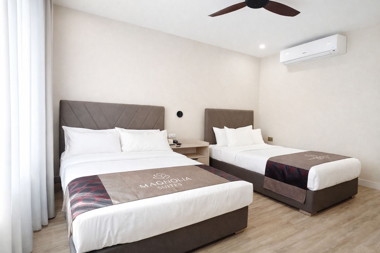 A hotel room with two beds, a ceiling fan, an air conditioning unit, and a nightstand with a lamp.