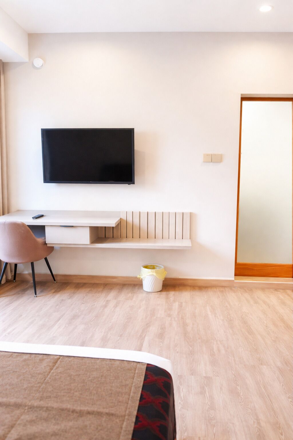 A modern hotel room with a flat-screen TV mounted on the wall, a wooden desk, a chair, and a bed with a patterned bedspread.