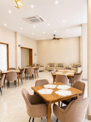 Un comedor amplio con varias mesas y sillas, un ventilador de techo, iluminación empotrada y un ventanal grande.