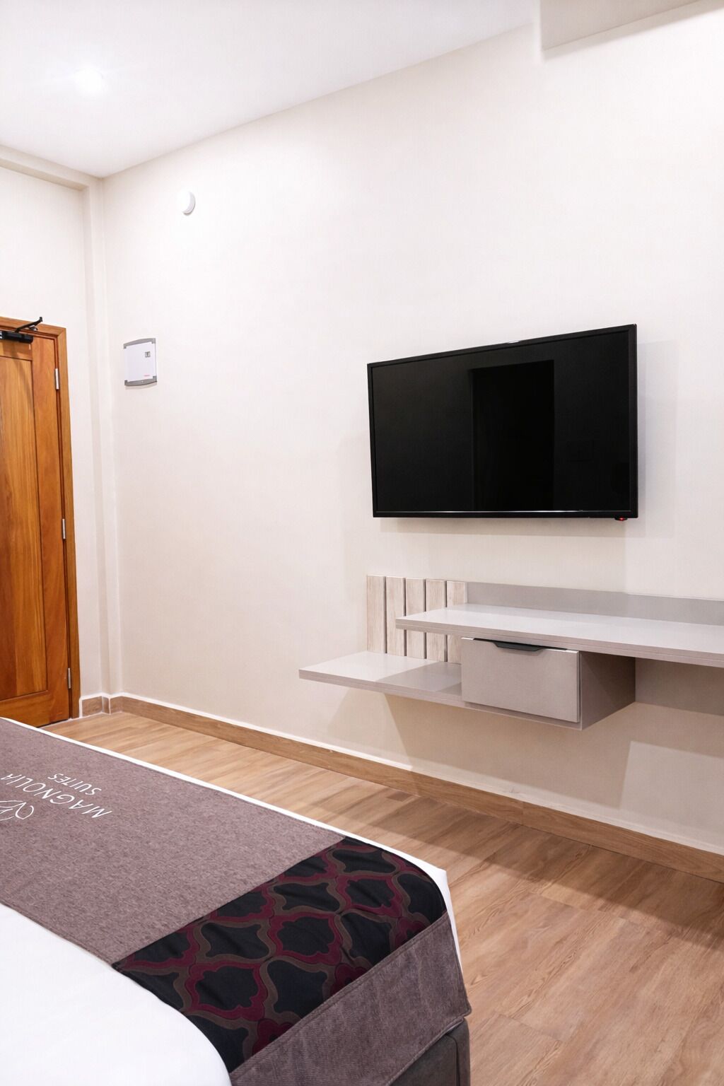 A hotel room with a flat-screen TV mounted on the wall, a wooden door, and a small shelf with a drawer.
