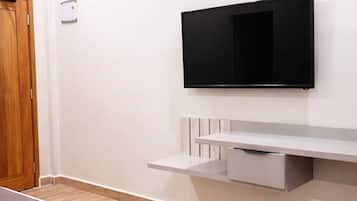 A hotel room with a flat-screen TV mounted on the wall, a wooden door, and a small shelf with a drawer.
