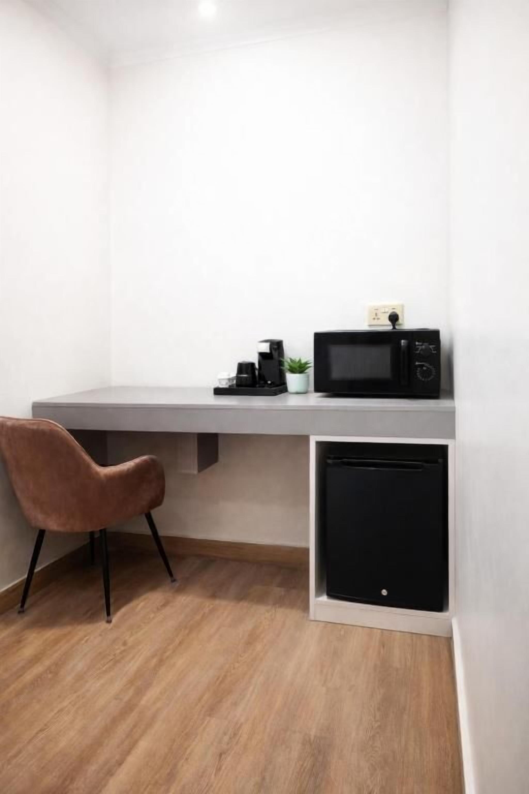 Una habitación compacta con un escritorio blanco, un microondas negro, un pequeño electrodoméstico negro y una silla marrón.