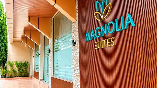 A hotel entrance with a wooden facade, a logo with a stylized flower, and the name 'Magnolia Suites'.