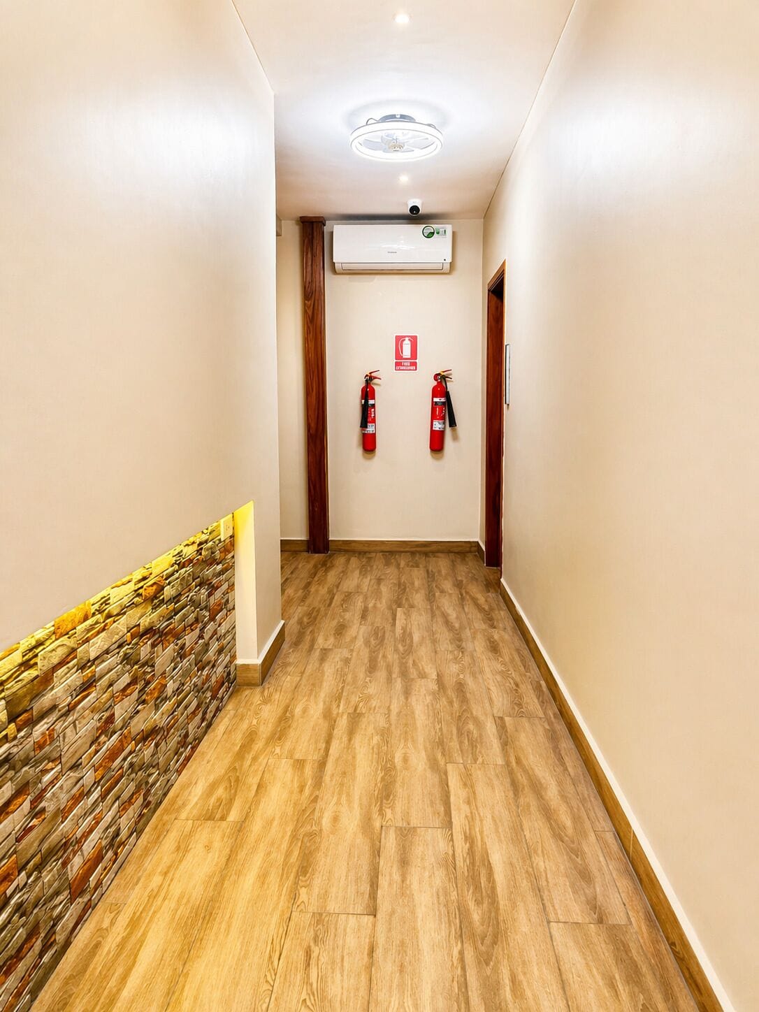 Un pasillo con suelo de madera, una pared de acento de piedra, una unidad de aire acondicionado y dos extintores montados en la pared.