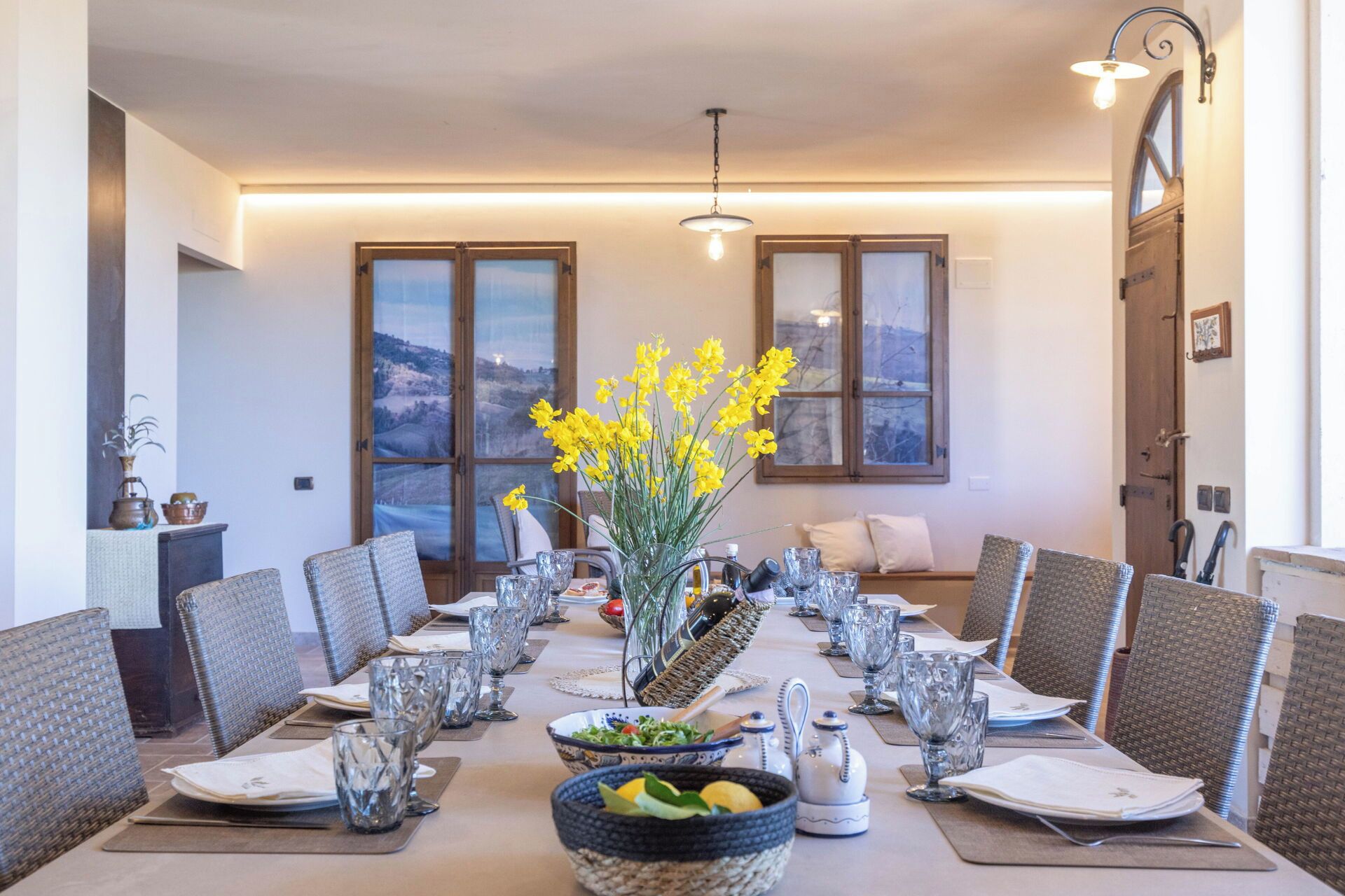 A dining area with a long table set for a meal, surrounded by wicker chairs, with a vase of yellow flowers in the center.