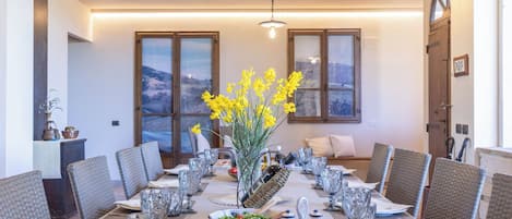 Un comedor con una larga mesa dispuesta para una comida, rodeada de sillas de mimbre, con un jarrón de flores amarillas en el centro.