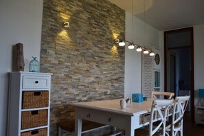 A dining area with a stone wall, a white table, wicker baskets, and white chairs. - Inviting Apartment With Pool Near the Beach (Lignano Sabbiadoro)