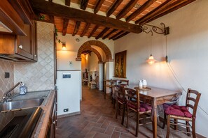 Cocina con pileta, estufa y muebles bajos de madera. Hay una zona de comedor con mesa y sillas, y un pasillo que conduce a otra habitación.