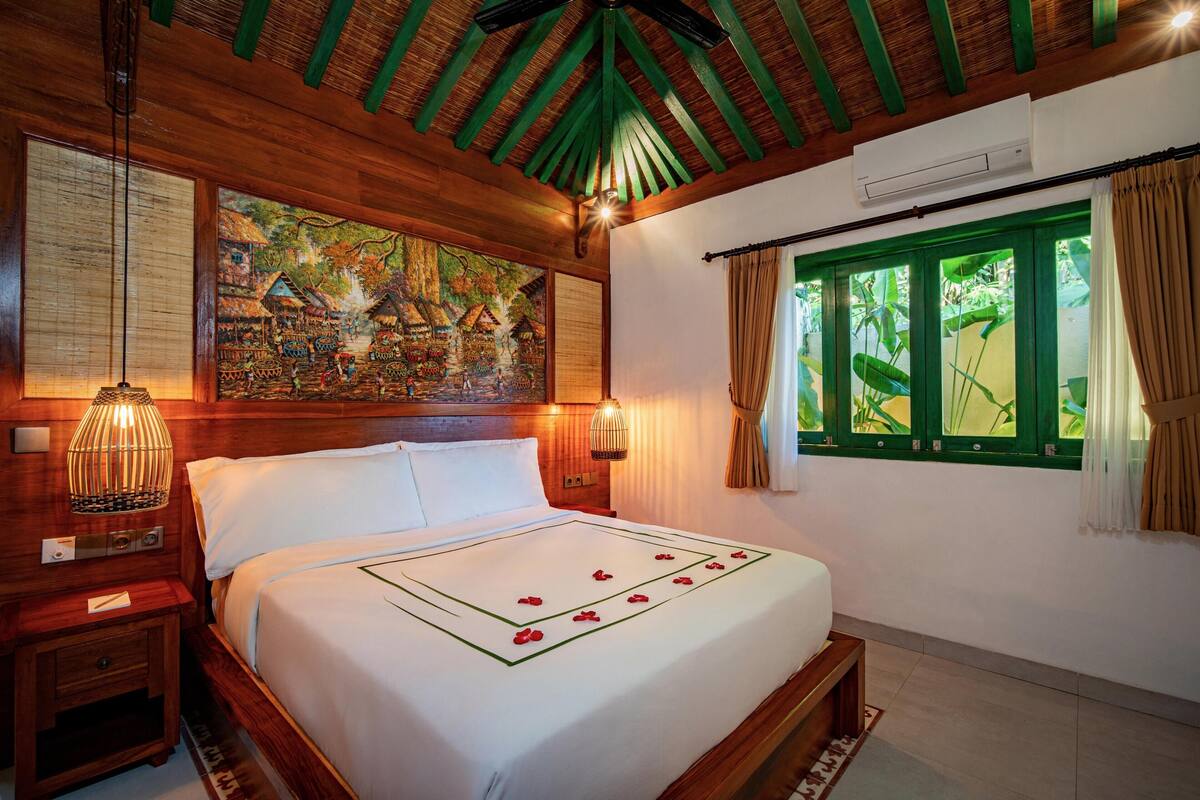 A wooden-framed room with a bed, a painting, a window with curtains, and a wall-mounted air conditioner.