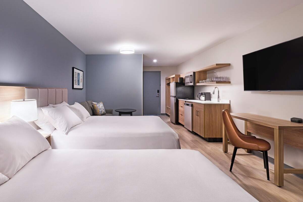 Ein Hotelzimmer mit zwei Betten, einem Schreibtisch, einem Stuhl, einem Fernseher und einer Küchenzeile.