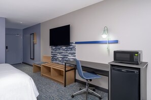 Una habitación de hotel con cama, escritorio con silla, televisor, microondas y refrigerador pequeño.