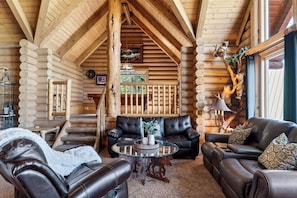 A log cabin interior with a fireplace, leather sofas, a glass coffee table, and a wooden staircase. - Hot Tub + Pool Table + Views | Bear Mountain + Lake | Moonshadows Log Cabin (Big Bear Lake)