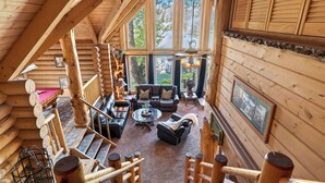 L’intérieur d’une cabane en rondins comprend une chambre à coucher située en mezzanine, un espace de vie avec une cheminée et de grandes fenêtres.