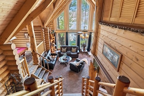 A log cabin interior with a loft bedroom, a living area with a fireplace, and large windows. - Hot Tub + Pool Table + Views | Bear Mountain + Lake | Moonshadows Log Cabin (Big Bear Lake)