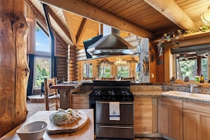 A log cabin kitchen with a stove, oven, and large windows. - Hot Tub + Pool Table + Views | Bear Mountain + Lake | Moonshadows Log Cabin (Big Bear Lake)