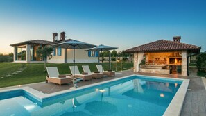 A swimming pool with lounge chairs and umbrellas, a stone fireplace, and a two-story house with a tiled roof. - Villa Evelyn by Villas Guide (Fažana)