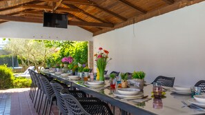 An outdoor dining area with a long table set for a meal, surrounded by chairs, with a TV mounted on the ceiling. - Casa Luce by Villas Guide (Pula)