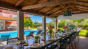 Ein überdachter Essbereich im Freien mit Pool, Liegestühlen und einem für eine Mahlzeit gedecktem Tisch.