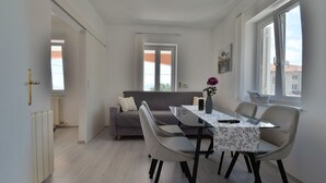 Een moderne woonkamer met een eettafel, bank, stoelen, een vaas met bloemen en grote ramen.