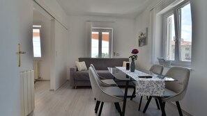 Ein modernes Wohnzimmer mit Esstisch, Sofa, Stühlen, einer Blumenvase und großen Fenstern.