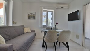 Een moderne woonkamer met een grijze bank, twee stoelen, een eettafel met een vaas en een aan de muur bevestigde televisie.