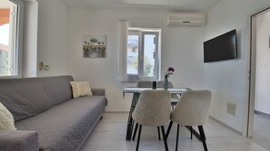 Ein modernes Wohnzimmer mit einem grauen Sofa, zwei Sesseln, einem Esstisch mit einer Vase und einem an der Wand befestigten Fernseher.