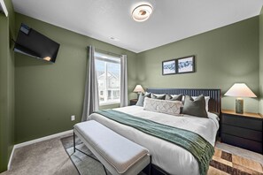 Ein Schlafzimmer mit Bett, Nachttischen, einer Bank, einem an der Wand montierten Fernseher und einem Fenster mit Vorhängen.