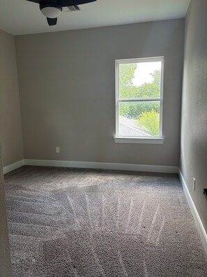Ein Zimmer mit Teppichboden, einem Fenster mit Blick auf Grünflächen und einem Deckenventilator.