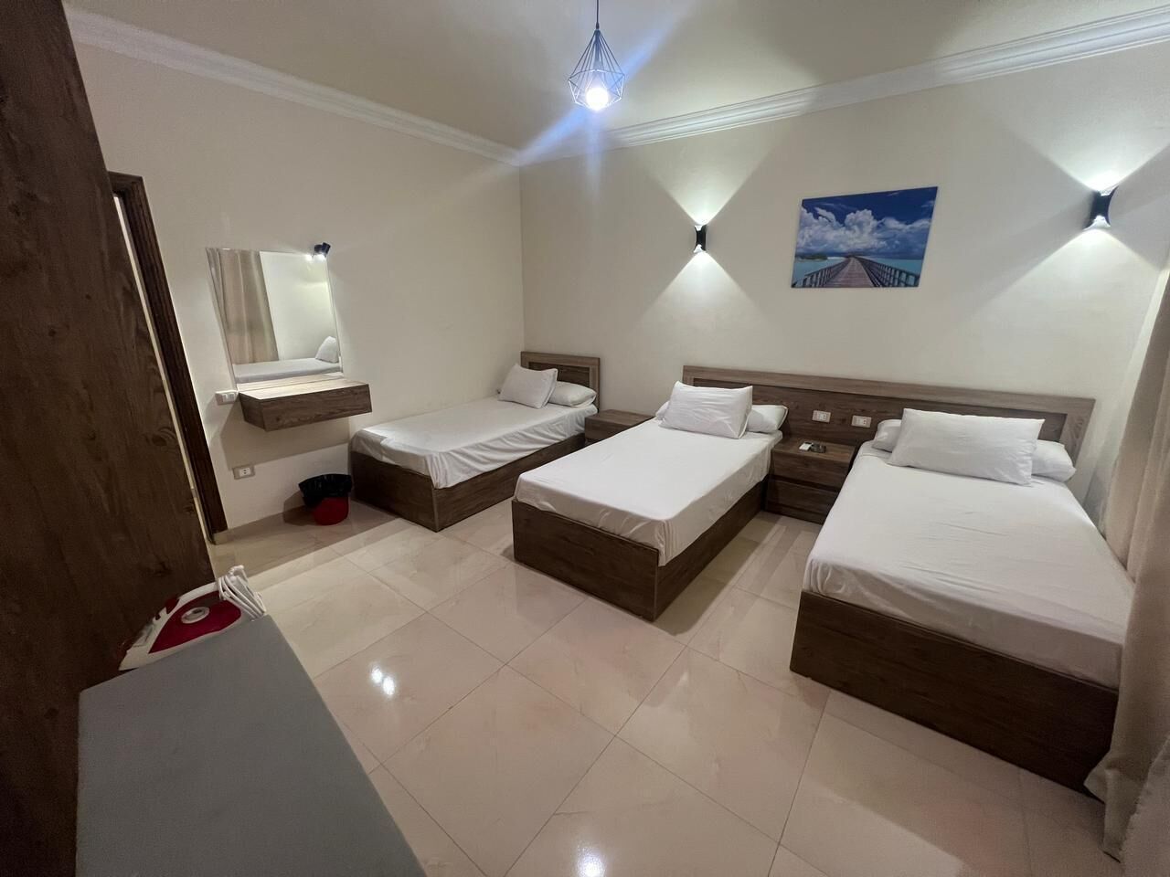 A hotel room with three beds, a large mirror, a picture on the wall, and a ceiling light.