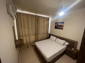Une chambre à coucher comprenant un lit, une table de chevet, un climatiseur fixé au mur, une fenêtre avec des rideaux et un tableau représentant un paysage urbain accroché au mur.