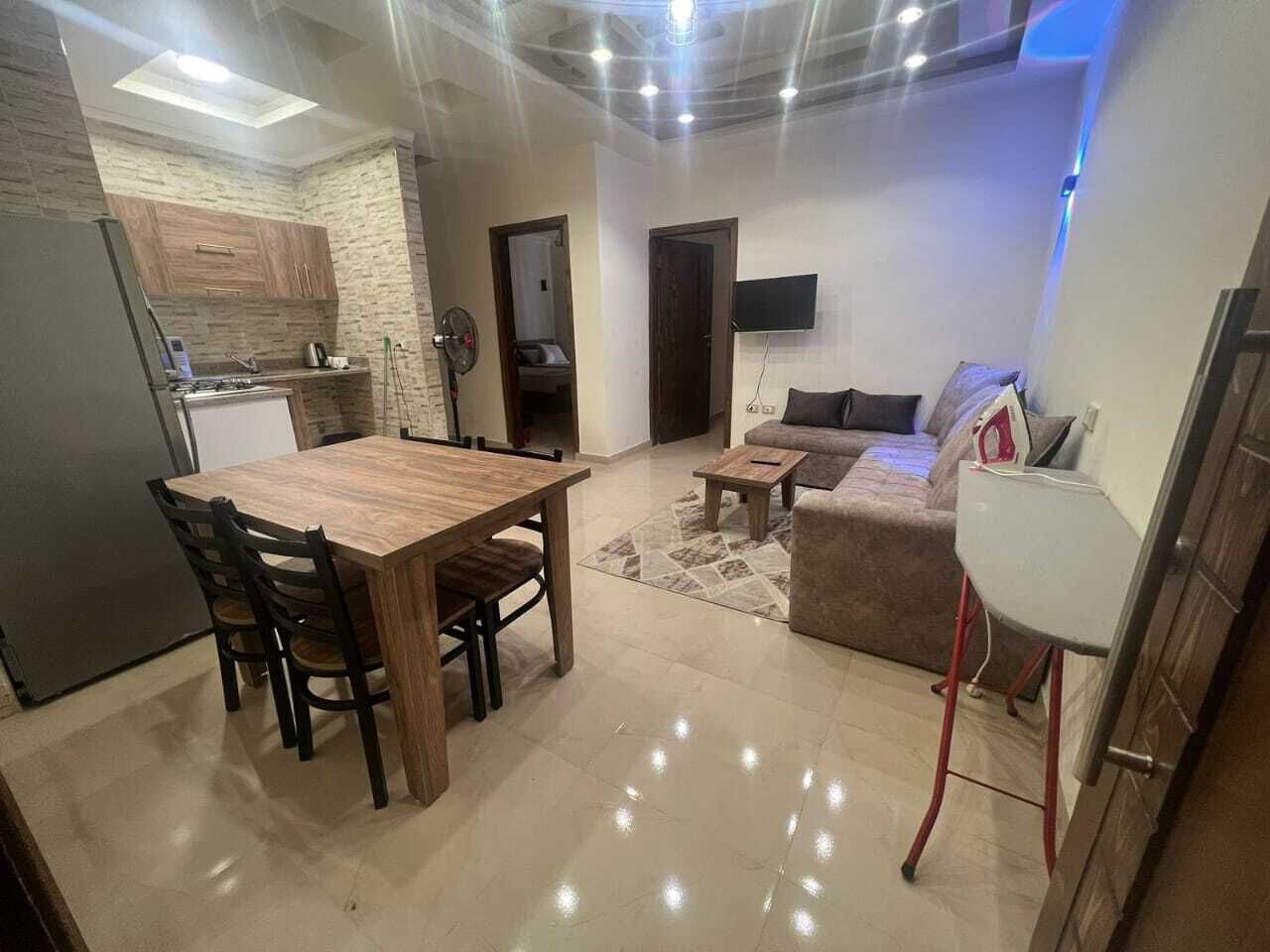 Una cucina moderna con forno integrato, un'area pranzo con tavolo e sedie in legno, un soggiorno con divano e tavolino, e una TV a parete.