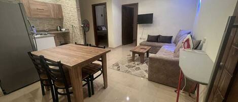A modern kitchen with a built-in oven, a dining area with a wooden table and chairs, a living room with a sofa and a coffee table, and a TV mounted on the wall.