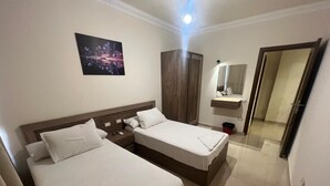 Habitación de hotel con dos camas, un armario de madera, un espejo y una obra de arte con un motivo urbano en la pared.