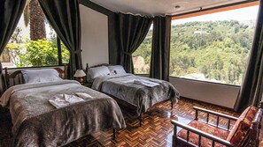 A room with two beds, a wooden floor, a view of a forest, and a chair.
