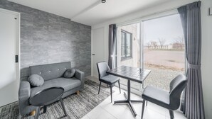 A modern room with a grey sofa, a small round table, two chairs, a large window with curtains, and a grey tiled floor. - ASTAGE CHITOSE SUEHIRO 103 (Chitose)