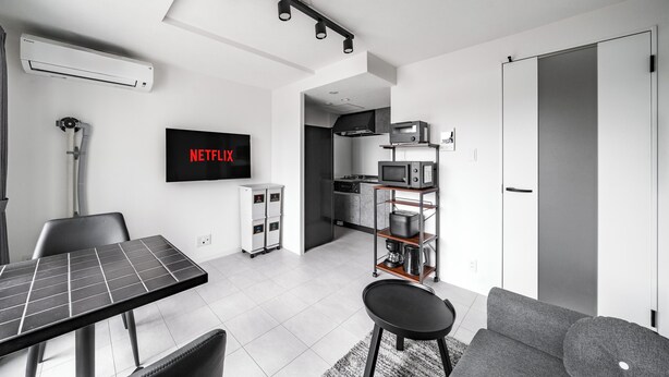 A modern living room with a flat-screen TV, a dining table, a sofa, and a kitchen area in the background. - ASTAGE CHITOSE SUEHIRO 103 (Chitose)