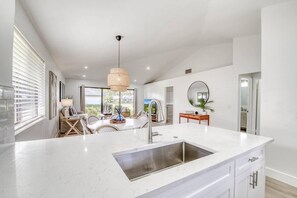 A modern kitchen with a large island, stainless steel sink, and a dining area with wicker chairs. - PGA National Cottage | Pool, Patio & Golf (Palm Beach Gardens)
