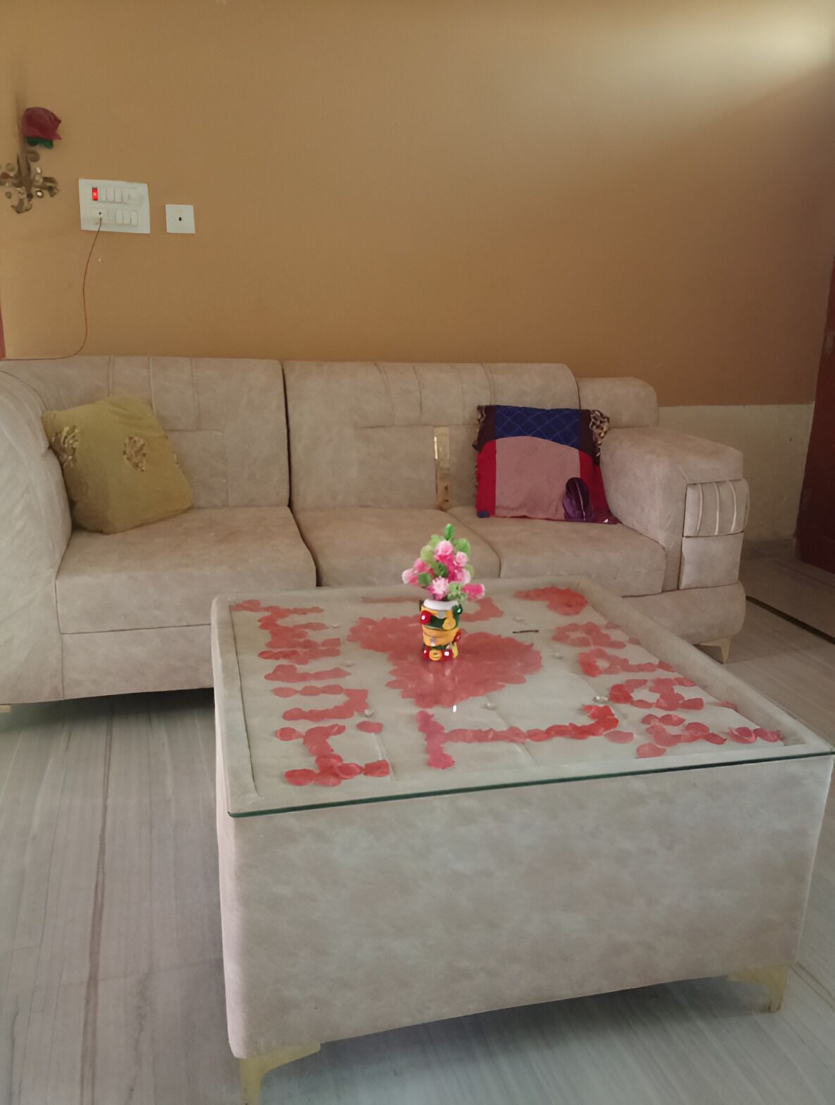 Un saló amb un sofà beix, una taula de centre de vidre amb pètals de rosa, un ram i una presa de corrent.