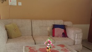 Una sala de estar con un sofá beige, una mesa de centro de vidrio con pétalos de rosa, un jarrón decorativo y un enchufe de pared.