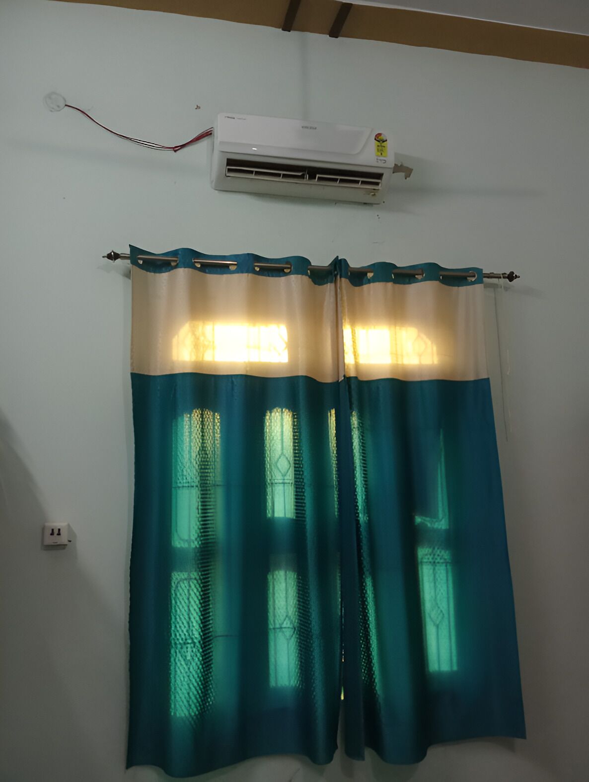 A room with a window, a curtain, an air conditioning unit, and a wall socket.
