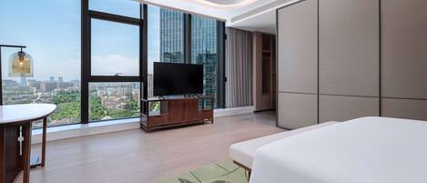 A modern hotel room with a large bed, a TV on a wooden stand, a dining table, and a view of the cityscape through large windows.