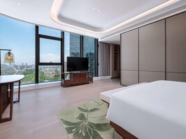 A modern hotel room with a large bed, a TV on a wooden stand, a dining table, and a view of the cityscape through large windows.