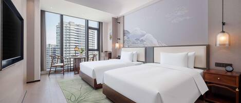 A hotel room with two beds, a large window, a flat-screen TV, a dining area, and a wall-mounted clock.
