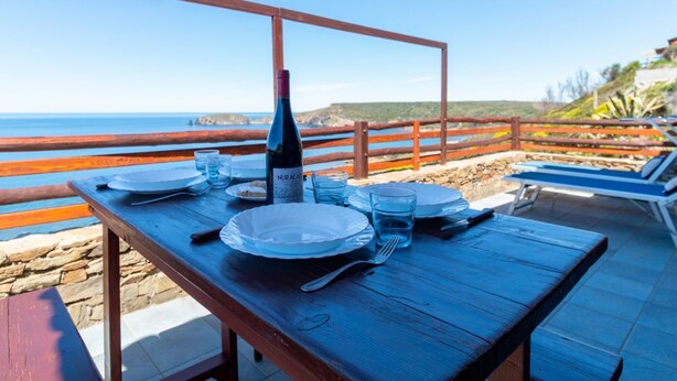 A wooden table set for dining with a bottle of wine, plates, and glasses, overlooking a beach with a wooden railing. - Casa Libeccio - Pistis - wifi - Pet Friendly (Arbus)