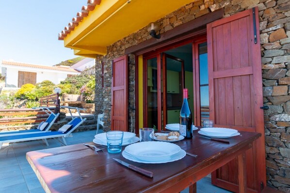 A wooden table with plates, a glass, and a bottle, set against a stone wall with red shutters. - Casa Libeccio - Pistis - wifi - Pet Friendly (Arbus)