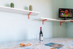 A room with a table, two white shelves with red brackets, a television, a bottle of wine, two glasses, and a plate with cookies. - Casa Libeccio - Pistis - wifi - Pet Friendly (Arbus)