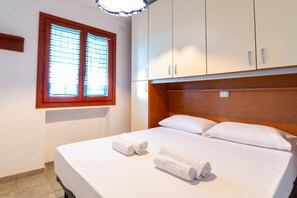 A hotel room with a bed, two rolled towels, a window with blinds, and wooden cabinets. - Casa Libeccio - Pistis - wifi - Pet Friendly (Arbus)