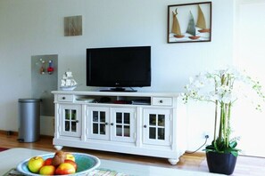 Ein Wohnzimmer mit einem weißen Schrank, einem Fernseher, einer Topfpflanze und einer Obstschale.