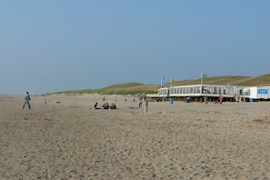 Ein Strand mit einem Gebäude, das wie ein Restaurant oder Café wirkt, mit Außenbestuhlung und einer Flagge.